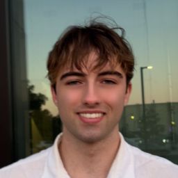 A person with short brown hair that is slightly messy wearing a white collared shirt looking directly at the camera and smiling.