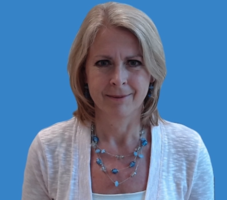 A female with slightly wavy shoulder length blond hair, wearing a white cardigan, a white top, and a three tiered necklace with large blue beads. She is looking directly at the camera with a slight smile.