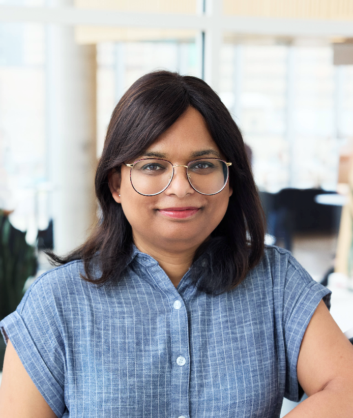 Profile of a brown woman in glasses with slight smile wearing a steel-coloured dress.