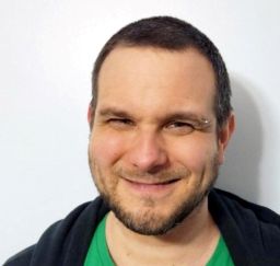 A white male with a buzz cut brown hair and a scruffy beard and mustache. He has a large smile on his face and is looking directly at the camera. 
