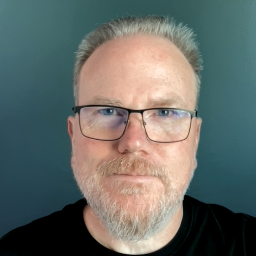 A person with a long buzzcut of blond and grey hair and a beard and mustache. He is looking directly at the camera, unsmiling. 