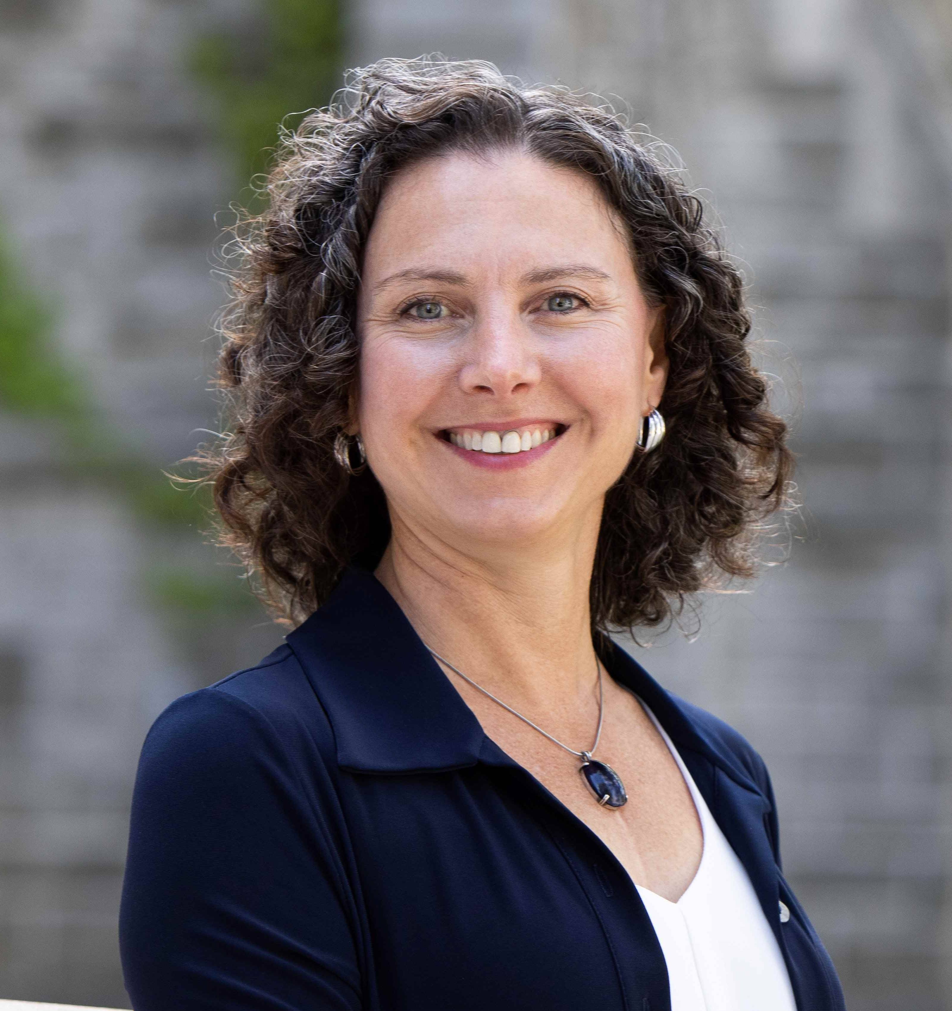 A white woman with shoulder length wavy brown hair. She is wearing a white top and a dark blue blazer. She is smiling widely at the camera.