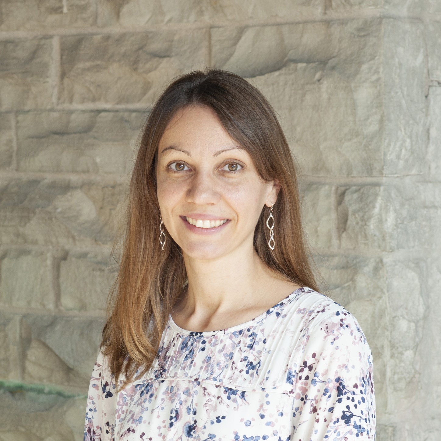 A white female with brown eyes and light brown hair past her shoulders, wearing a flowered blouse and spiraled earrings. She is smiling and looking directly at the camera.