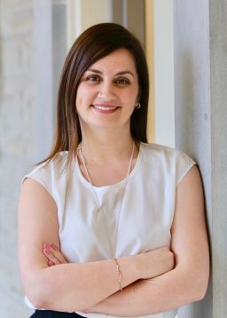 A female with shoulder length brown hair and light skin wearing a white short sleeved top. She is looking directly at the camera and smiling. 