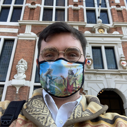 A male with short brown hair wearing round wire-rimmed glasses and a large face mask covering the bottom half of his face. He is standing in front of a red brick building and looking directly at the camera.