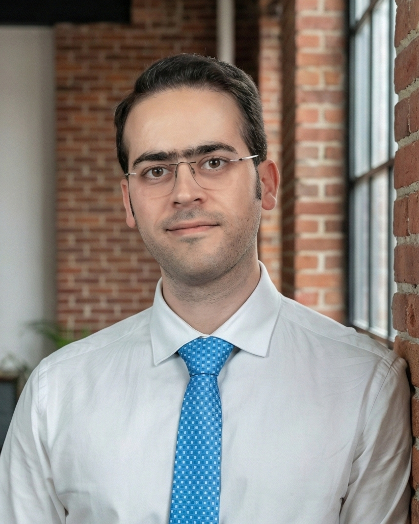 A portrait of a male with short dark hair, light stubble, and rimless glasses, wearing a white shirt and blue tie, leaning against a red brick wall next to a large industrial window, with a slight, confident smile. 