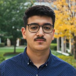 A brown male with short black hair, a mustache, and a slight goatee wearing a blue collared shirt and black glasses. He is looking directly at the camera, unsmiling.