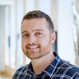 A person with short curly brown hair and a short beard and mustache. He is wearing a blue checkered collared shirt. He is looking directly at the camera with a slight smile. 