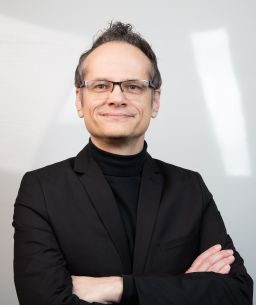 A white male with short brown hair and square glasses wearing a black turtleneck and blazer. He looking directly at the camera with a smile and arms crossed.