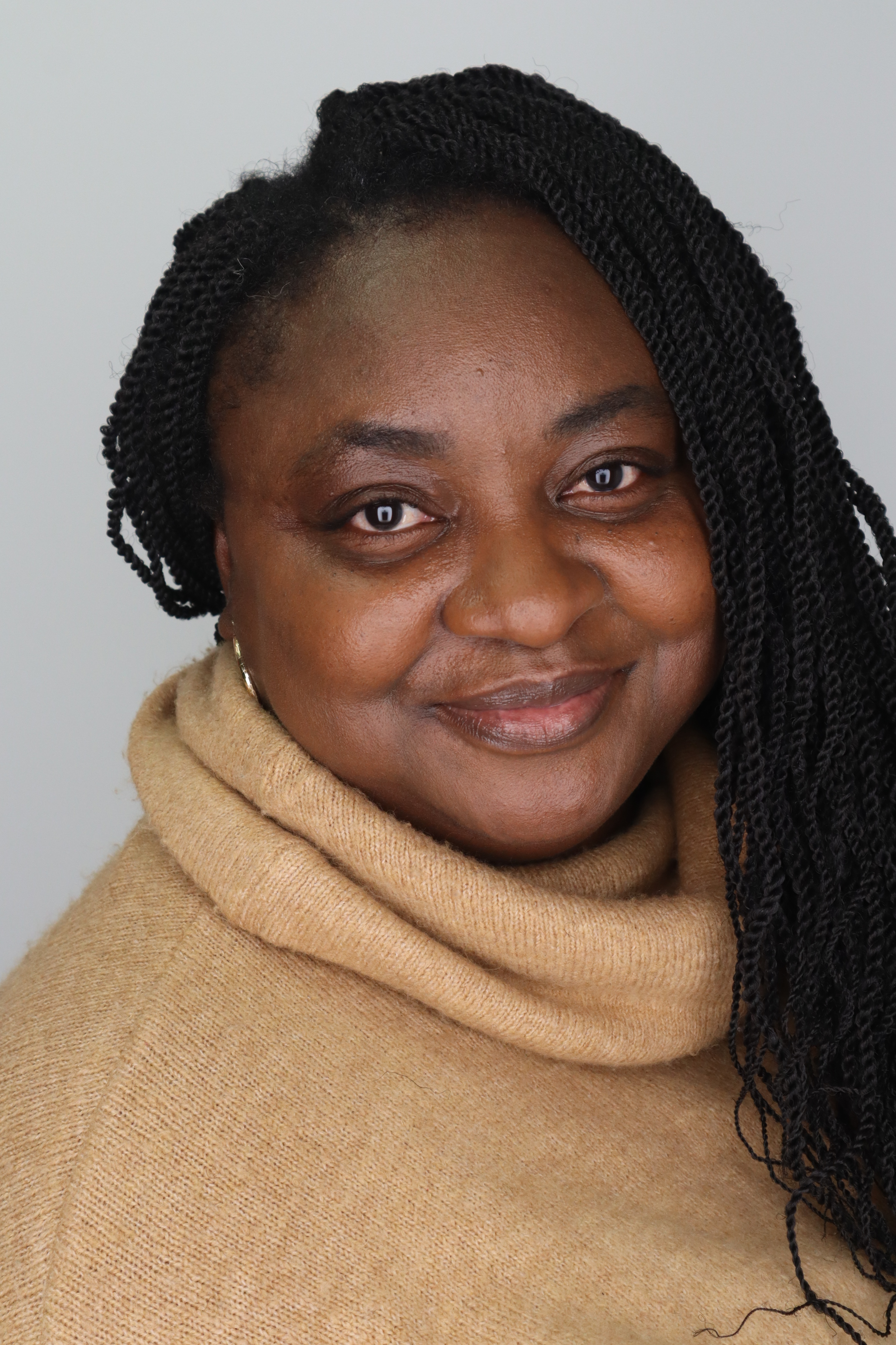 A portrait of a medium-dark brown skin woman with long, twisted braids, wearing a soft, camel-coloured turtleneck sweater. The background is a plain light grey, and the lighting is even, highlighting their warm, friendly expression.