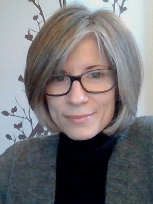 A female with straight silvery-grey shoulder length hair, wearing glasses, a black top and a grey blazer. She is looking at the camera with a slight smile.  