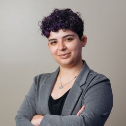 A person with short curly purple hair wearing a grey blazer with their arms crossed. They are looking at the camera with a slight smile. 