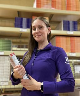 A white female with long brown hair waring a purple jacket with a Western logo on the sleeve. She is holding a textbook in her hand. She is looking directly at the camera with a slight smile.