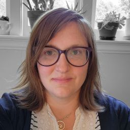 A white female with reddish brown shoulder-length hair. She is wearing over-sized glasses and a white and black striped shirt with a black blazer. She is looking directly at the camera with a slight smile. 