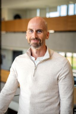 A bald white man with blue eyes and a short salt-and-pepper beard. He is wearing a cream quarter-zip sweater over a white shirt, looking directly at the camera with a slight smile.