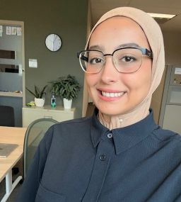 The image displays a selfie of a smiling woman wearing a beige hijab, glasses with black upper frames, and a dark button-down shirt. She is indoors in an office setting, with an olive-green wall, a round white wall clock, an office chair, and small potted plants resting on a cabinet visible in the background.
