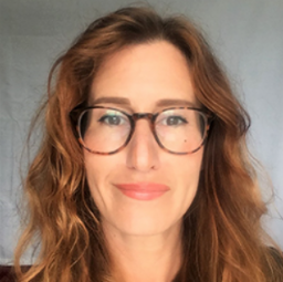 A white female with long wavy red hair. She is wearing glasses and looking directly at the camera with a slight smile.